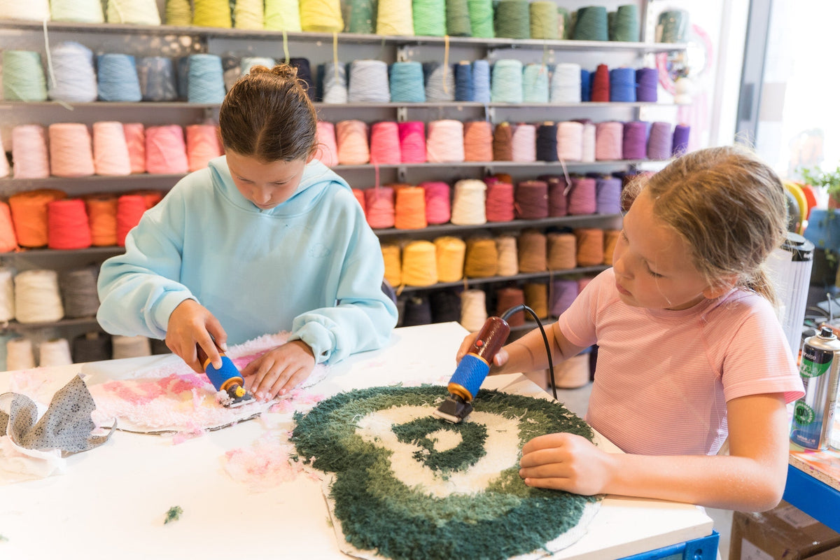 Two young girls tufting rugs at a worktable inside Cheeky Studio, Hackney Wick — colourful yarn cones displayed on shelves in the background