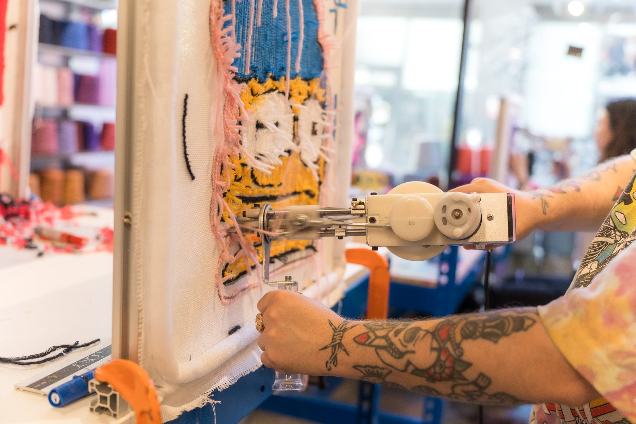 Close-up of a tufting gun being used to create a yellow and white smiley face design on a tufting frame, with yarn cones in the background