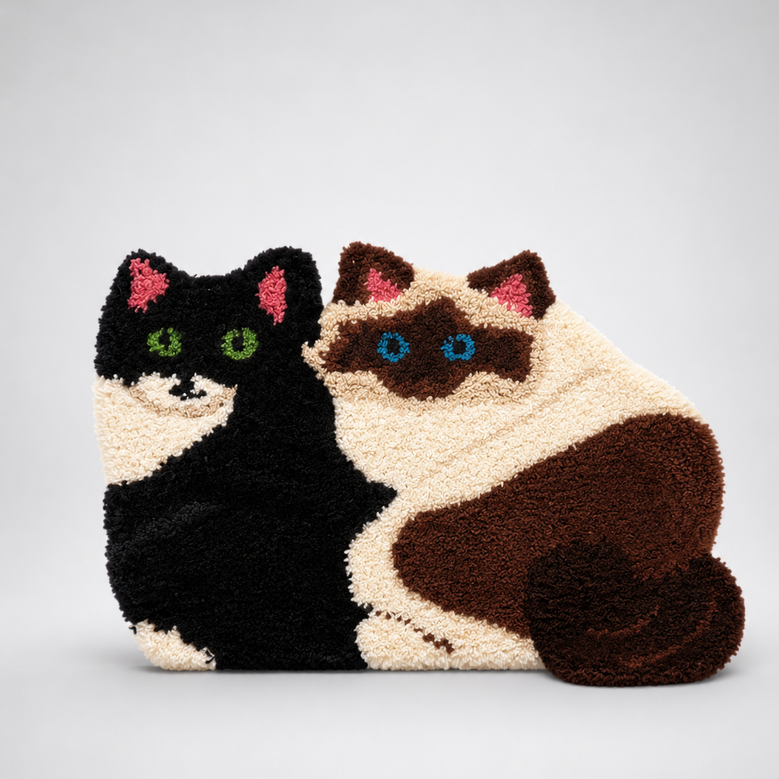 Tufted rug shaped like two sitting cats side by side — a black and white cat with green eyes and a Siamese-style cat with blue eyes, made using deep brown, cream, black, pink, and blue yarn